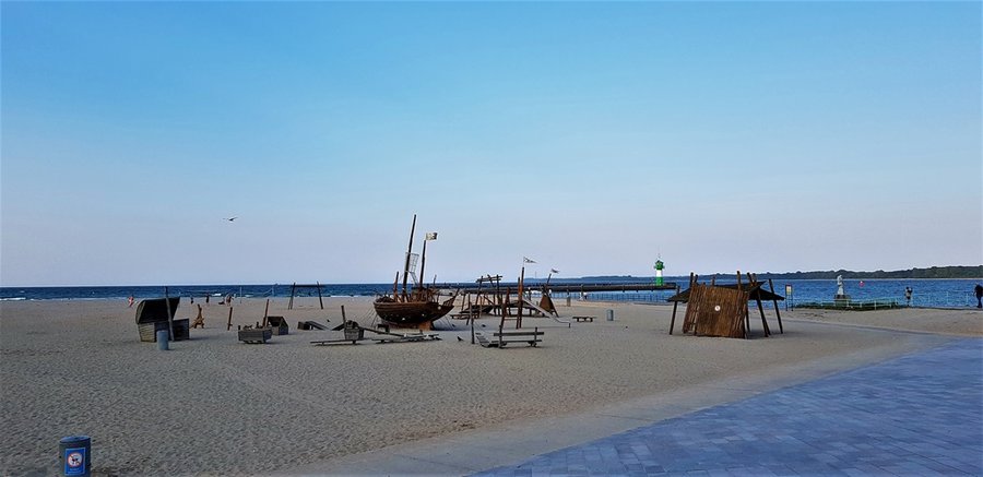 Piaszczysta plaża w Travemünde z drewnianym placem zabaw w kształcie żaglowca i zieloną latarnią morską w tle